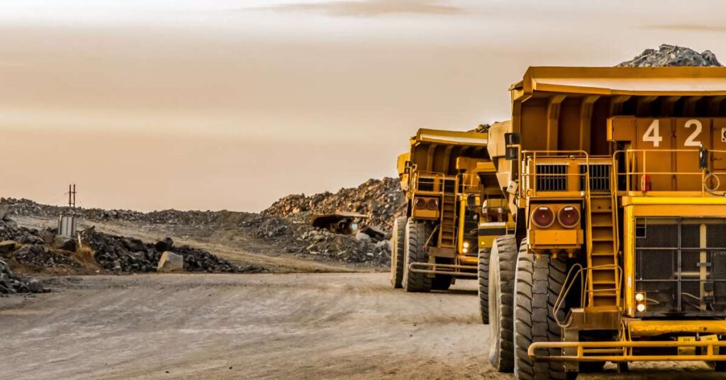 Two yellow mining dump trucks are driving around a platinum mine with dirt and debris in the background.