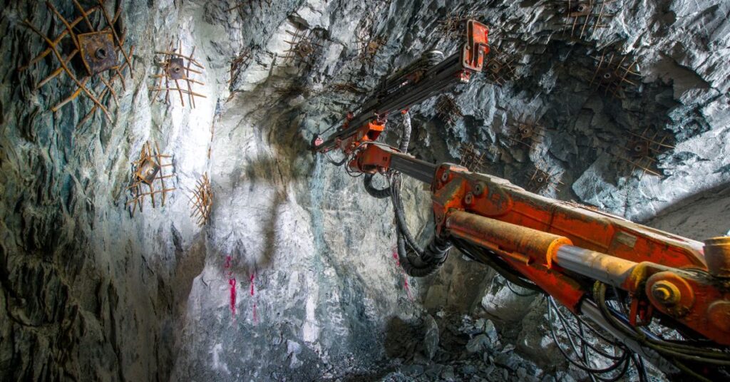 A large mining vehicle working underground in a hard rock mine. A large orange arm extends toward the mine wall.