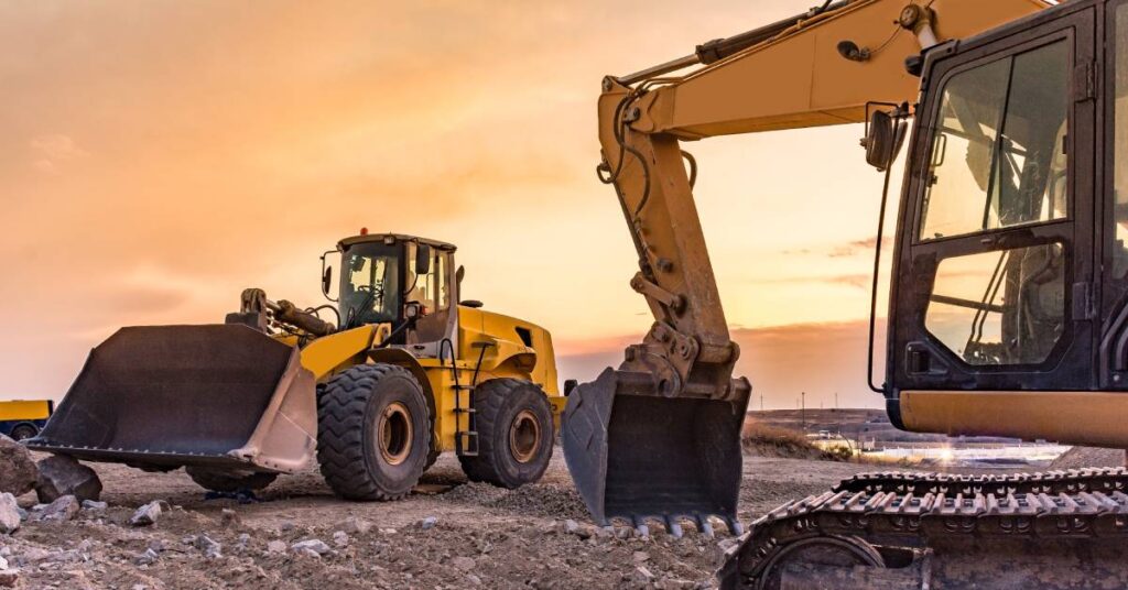 Two excavator heavy mining machines work together on a mining site. The sun and some clouds are behind them.