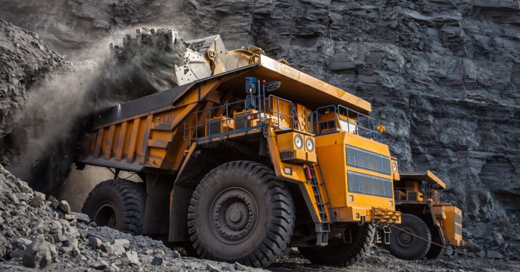 A massive yellow mining vehicle loads coal into the back with a wall of stone and another mining vehicle behind it.