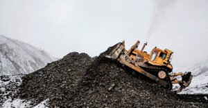 A bulldozer at a mining site climbs up on a hill of material while the background is full of clouds and snow.