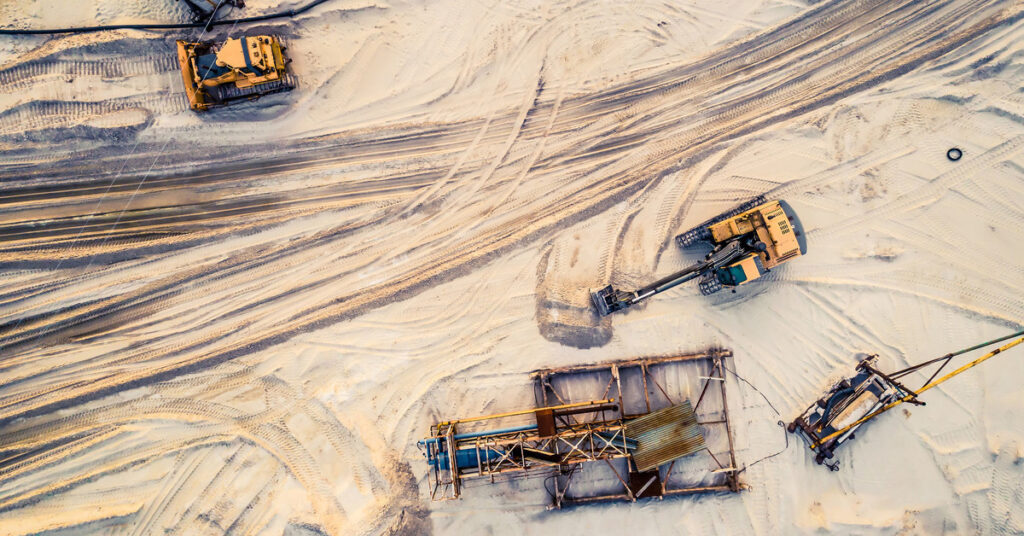 Different mining machines driving around in a sandy area. There are lines in the sand showing where machines are driving.