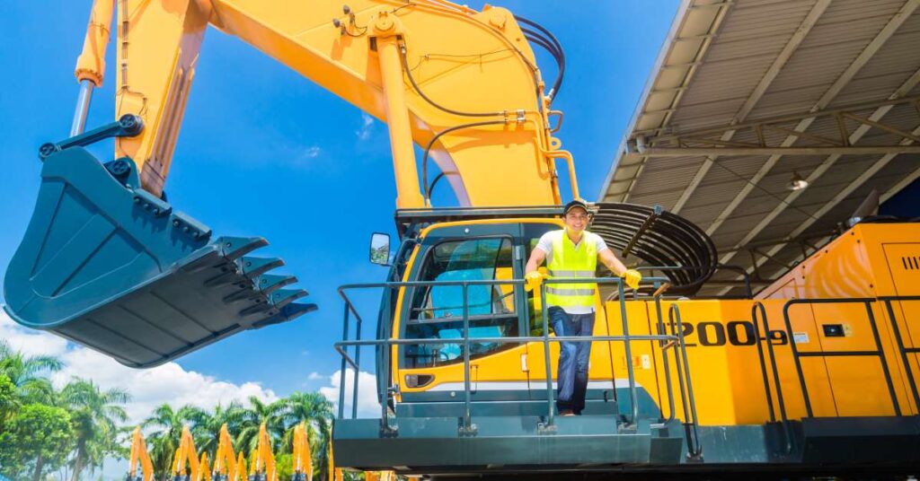 A worker in a hi-vis vest standing in front of a big yellow excavator and a bright blue sky on a construction site.