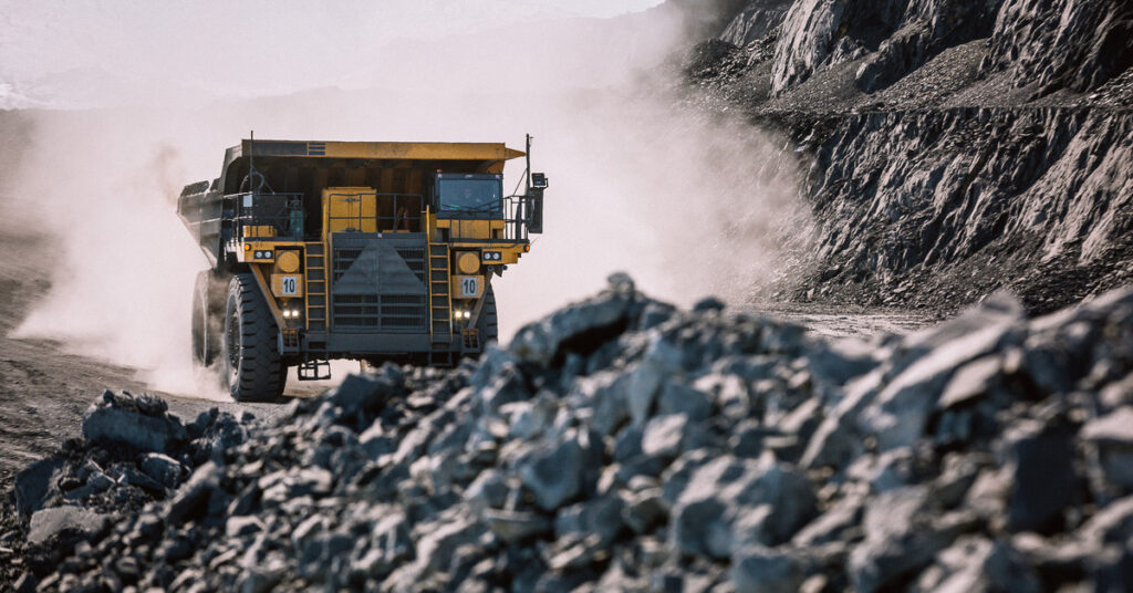 A dump truck drives down a mining site, kicking up clouds of smoke in its wake with lots of rocks in the foreground.