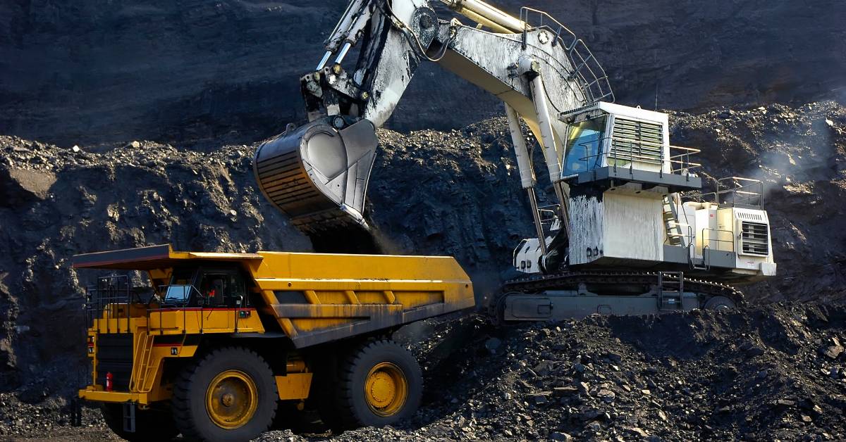 At a coal mining site, a white machine transfers coal and other rock into the back of another yellow mining vehicle.