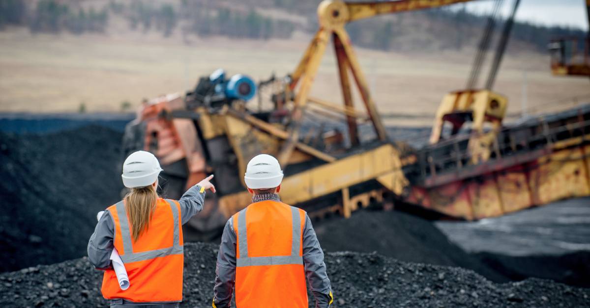 Two workers in orange hi-vis vests and hard hats supervise a coal mining site, pointing at a big mining machine.