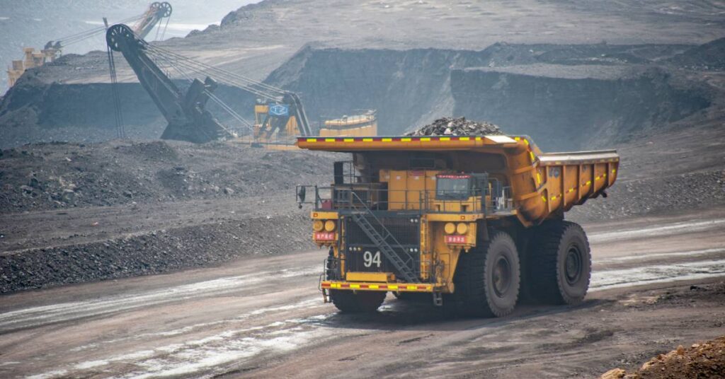 A large yellow mining truck hauls rocks in a coal mine. The mine is surrounded by dust with other machines in the background.