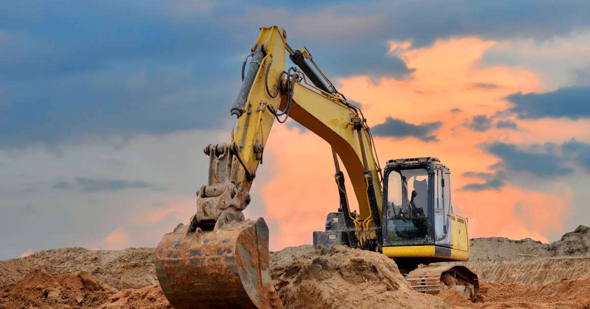 A large yellow excavator moves around big piles of dirt. In the background is a bright sunset peeking through the clouds.