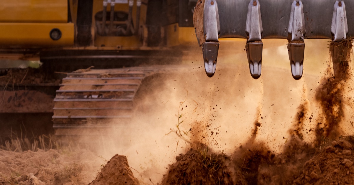 A close-up of a backhoe digging massive amounts of soil, kicking up dirt and dust with the bucket teeth.