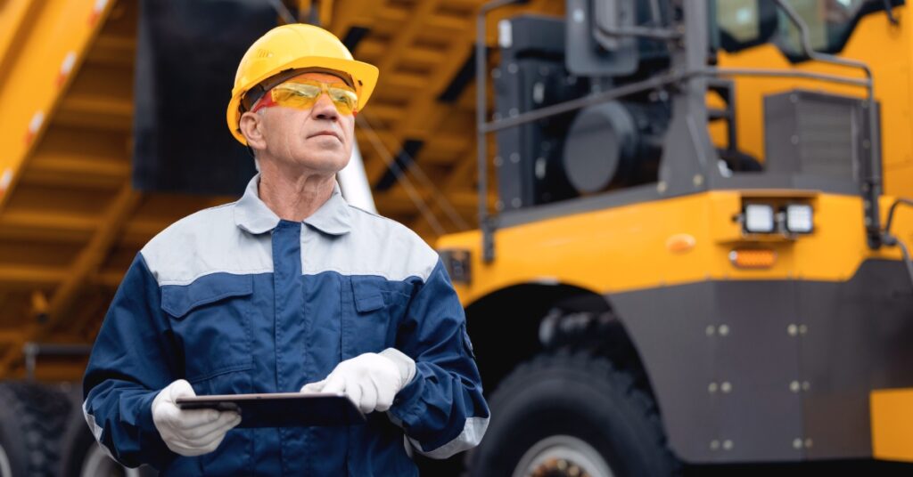 A mining operator wearing safety goggles and a yellow helmet holds a clipboard while surveying the mining site.