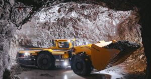 A large yellow underground mining digger picks up a big pile of mined material in a tight cave with little wiggle room.