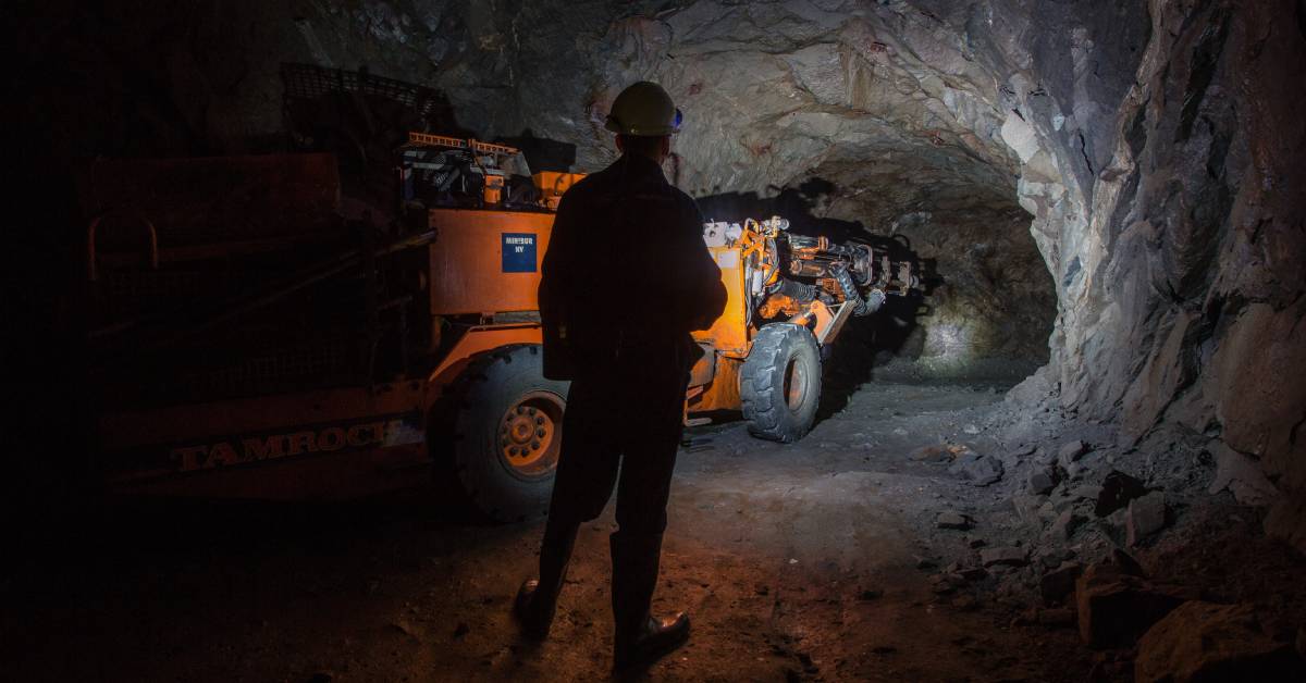 A dark mining tunnel with a worker standing in the dark, supervising an orange machine going further in.