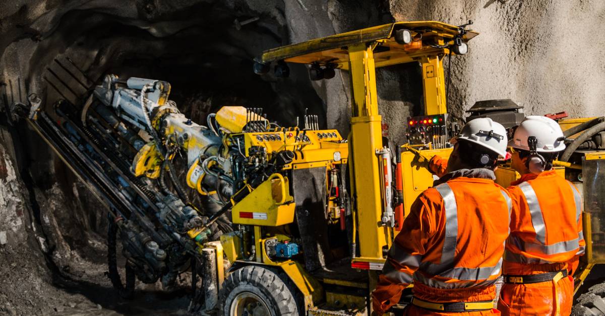 Two workers wearing orange work gear and helmets supervise a yellow mining machine drilling in an underground tunnel.