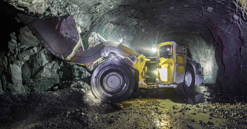 A large yellow mining vehicle operates in an underground hard rock mine with its lights illuminating the tunnel.