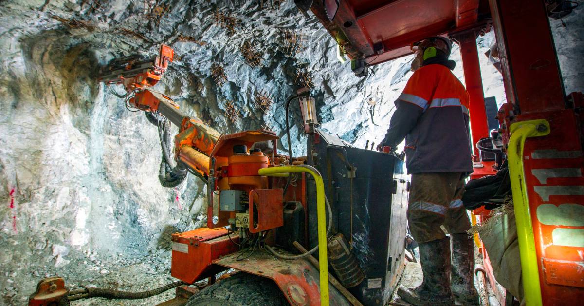 A worker stands on a large machine, pointing the machine's hydraulic arm toward the side of a tunnel.