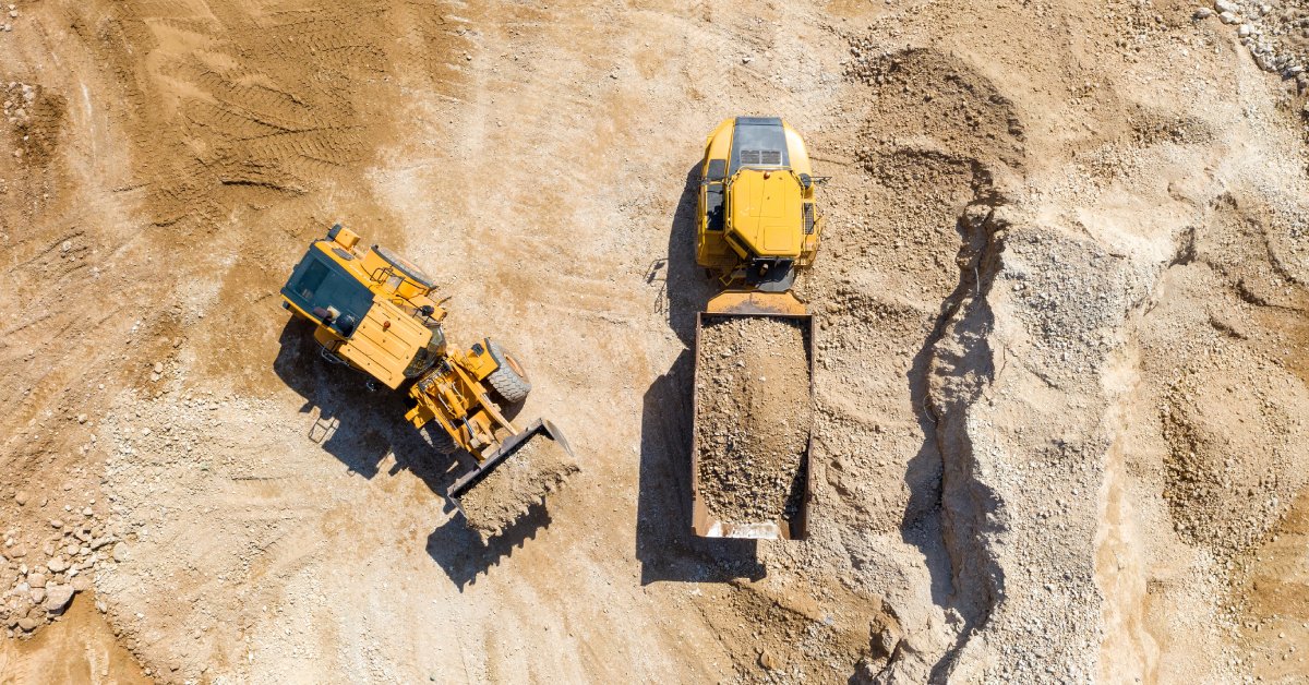 An aerial view of two mining vehicles with different attachments in a sandy area. Both are holding sand in their buckets.