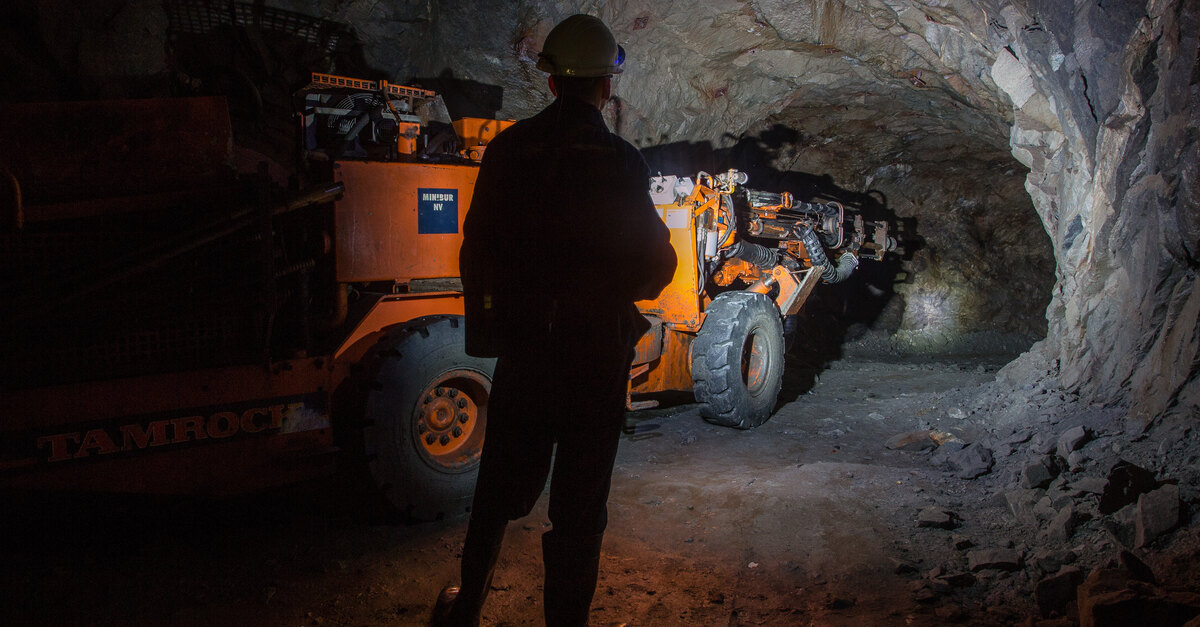 A miner supervises a yellow mining vehicle driving through a dark and cramped underground mining tunnel.