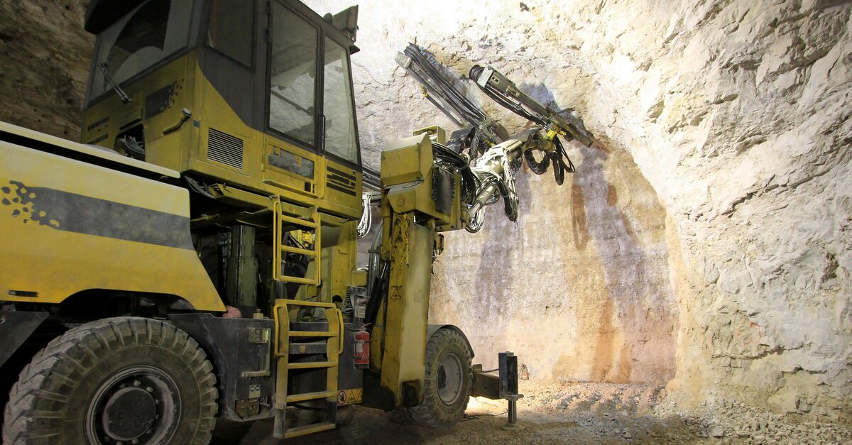 A large yellow mining vehicle works to clear some material off of a wall that a large light is illuminating.