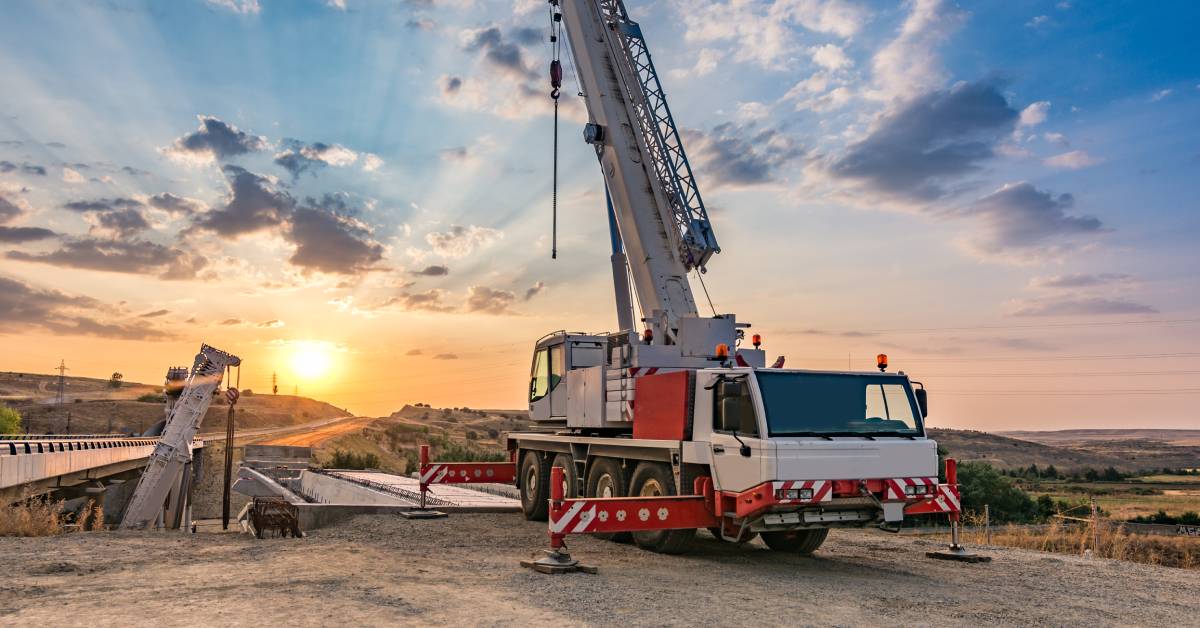 A mobile crane truck sits at a construction site featuring a partially finished bridge. The sun sets in the distance.