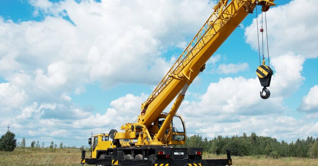 A mobile crane sits in a field with trees in the distance. The crane is large and yellow with a large hook dangling.