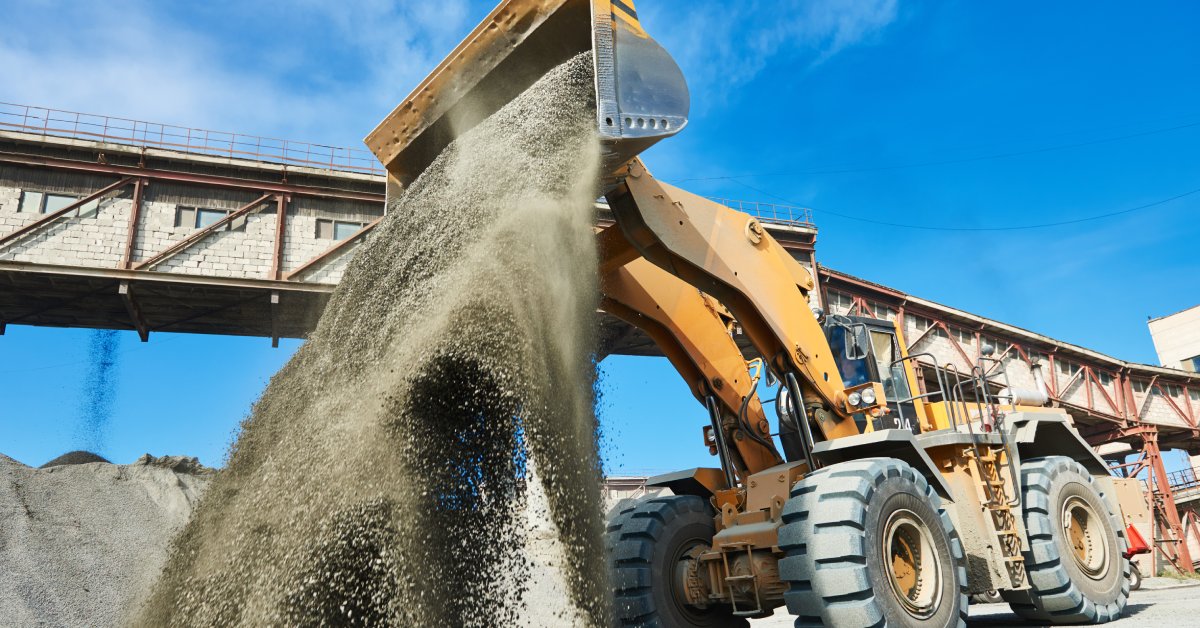 A mining vehicle dumping gravel and dirt from its bucket onto the ground. There is a long building behind the vehicle.