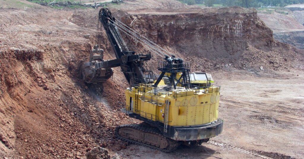 A large yellow mining vehicle with a crane shoveling dirt at the bottom of a deep trench that is halfway cleared out.