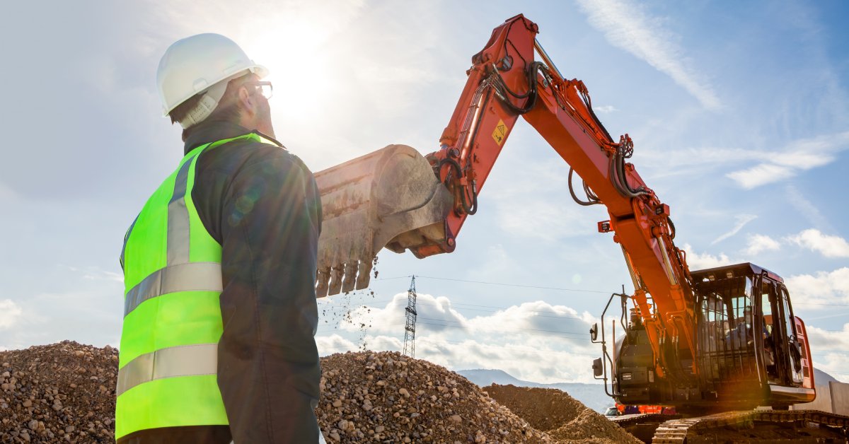 A worker standing in front of a mining excavator at a distance. He is wearing a white hard hat and a reflective vest.