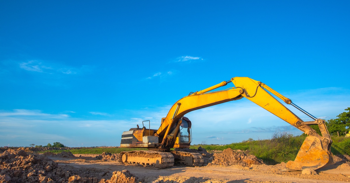 A yellow backhoe excavator parked on a flat road with the bucket at the end of the arm resting on the dirt.