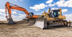 Two large yellow unattended mining vehicles are parked in a flattened dirt lot under a partly cloudy sky.