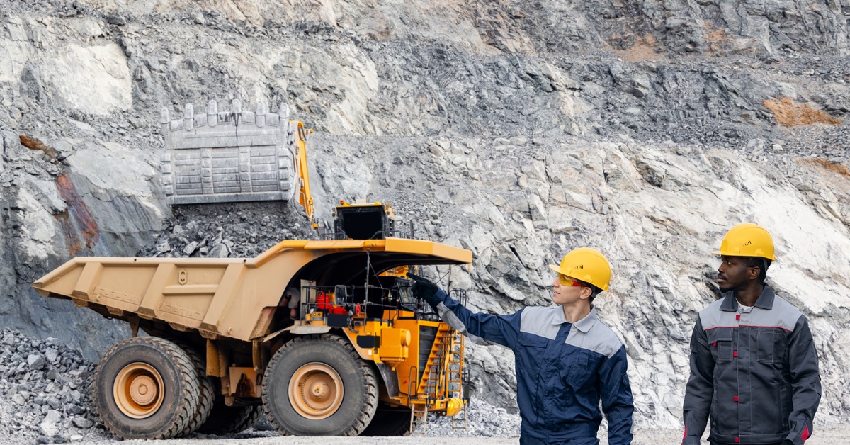 Two industrial workers wearing hard hats walk together outside with a large mining vehicle in the background.