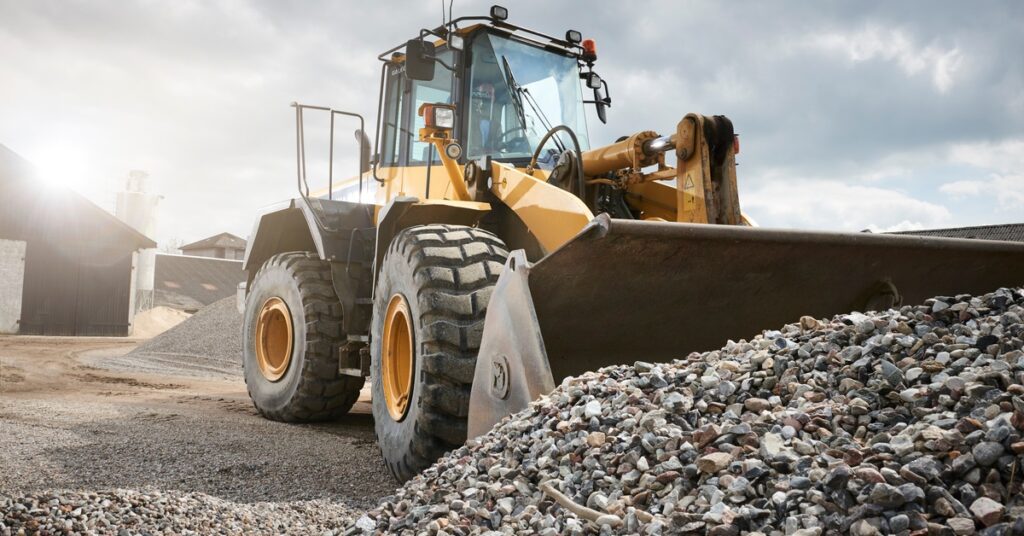 A mining front loader parked outdoors in a gravel lot. The bucket is near a large pile of gravel and rocks.