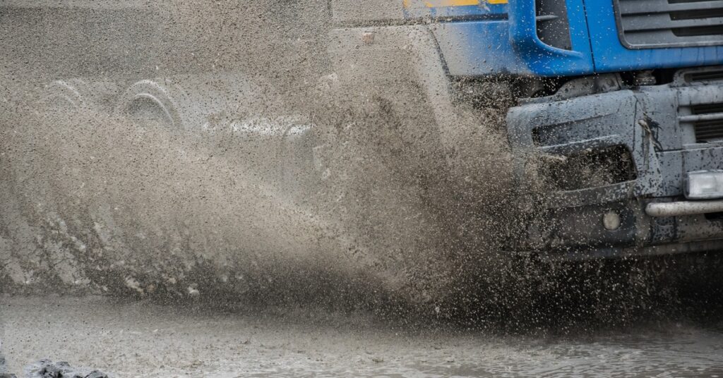 A blue industrial vehicle driving through a large puddle of muddy water and creating a large splash underneath the tires.