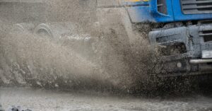 A blue industrial vehicle driving through a large puddle of muddy water and creating a large splash underneath the tires.