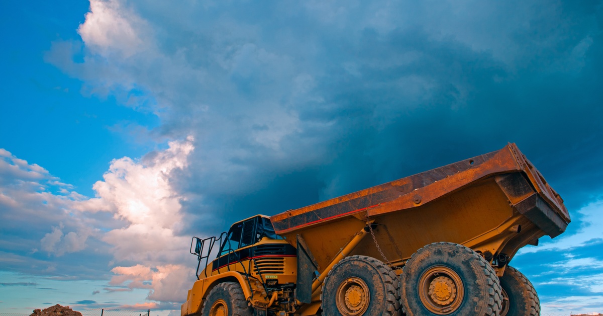 An orange mining truck parked outside on a dirt mound with large clouds partly covering the blue sky overhead.