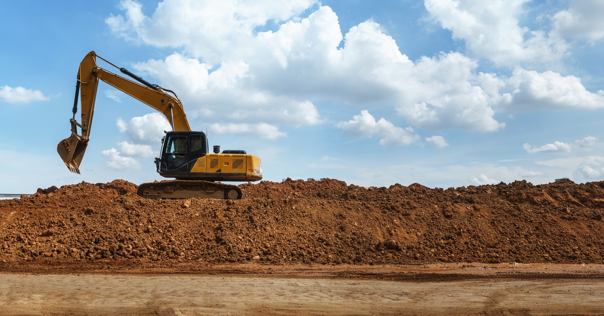A yellow excavator parked on top of a large mound of soft dirt. The sky behind the vehicle is partly cloudy.