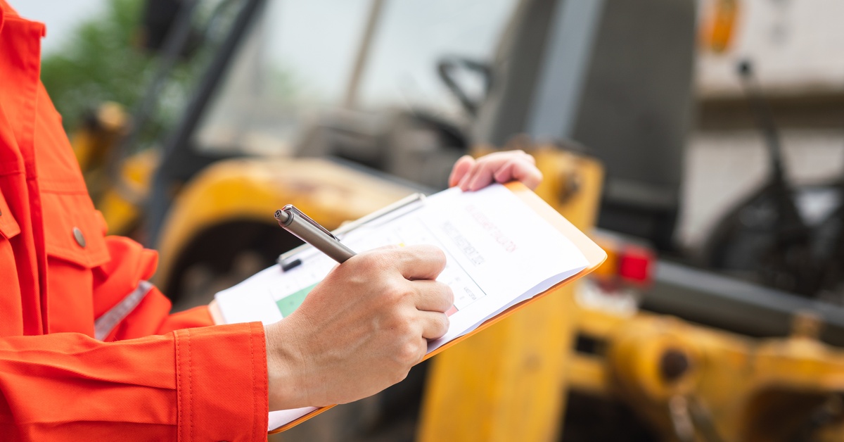 A person in an orange high-visibility jacket stands in front of a mining vehicle and writes on a clipboard.