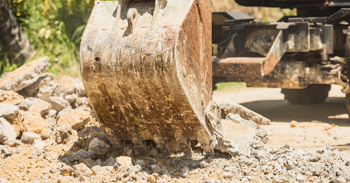 An excavator picking up a pile of rocks and gravel using its backhoe bucket. There are scuff marks and dirt on the bucket.