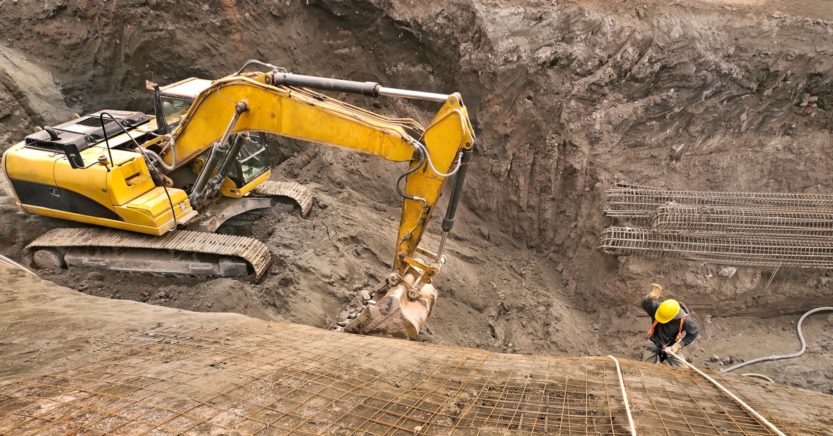 An excavator parked at the bottom of a dirt lot. There are site workers climbing and clearing the dirt paths and walls.