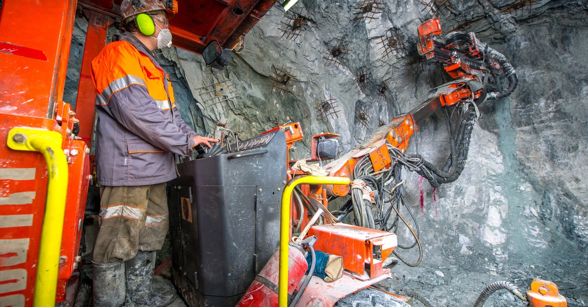 A person wearing ear protectors, goggles, and a mask operates an off-highway vehicle inside a mineshaft.