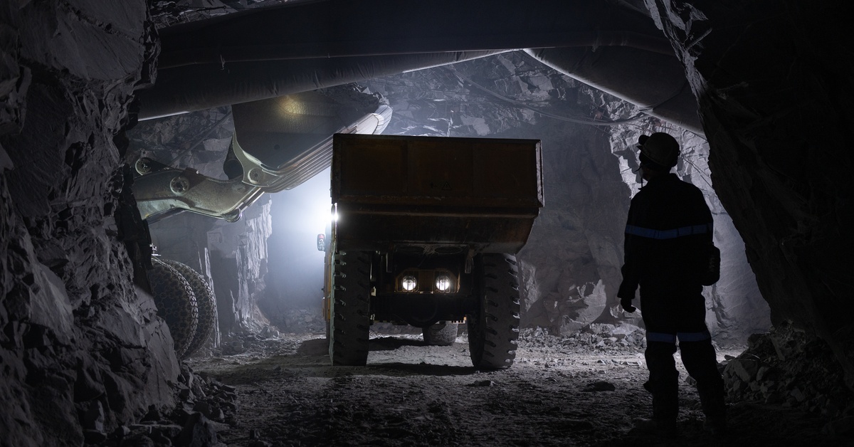 A person stands in one tunnel of a mine while watching two off-highway vehicles operate in front of them.
