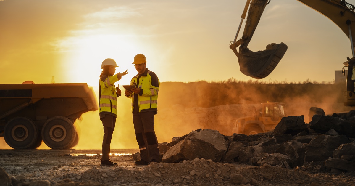 Two mining technicians wearing protective gear are talking in the middle of a worksite in between industrial vehicles.