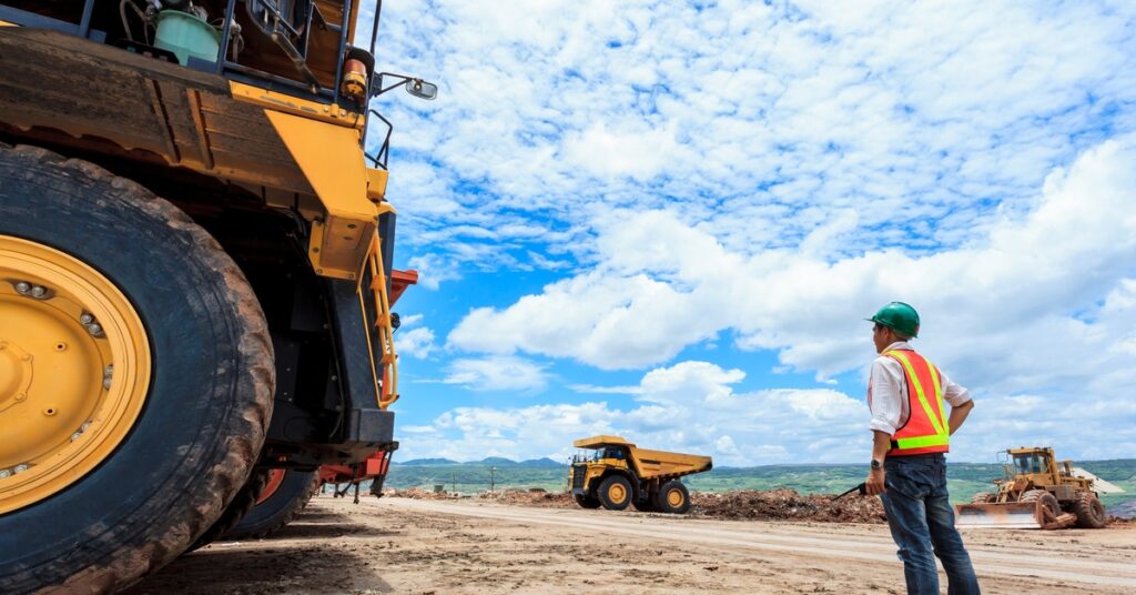 A worker wearing a high-visibility vest and helmet surveys an outdoor site with various mining vehicles in the distance.