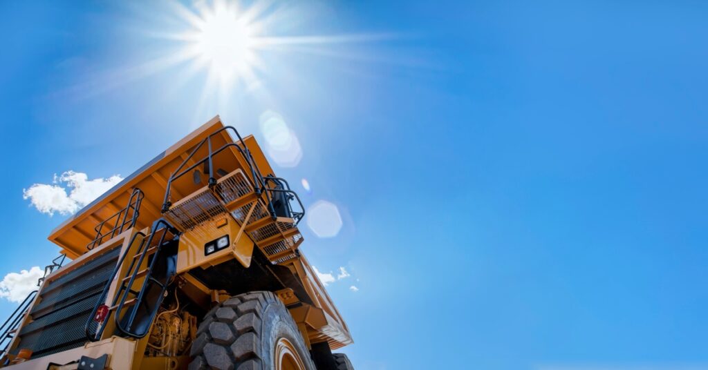 A large yellow mining vehicle sits still while the sun shines brightly above it with just a few clouds in the sky.