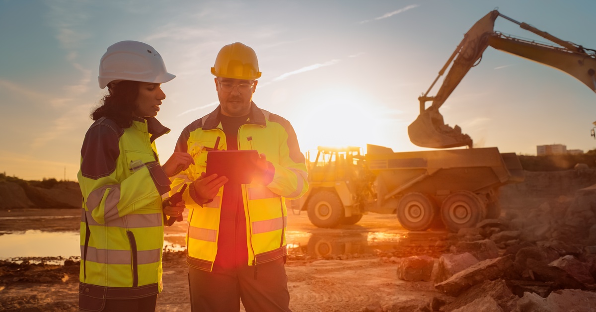 Two workers wearing helmets and high-visibility vests look over a clipboard with mining vehicles behind them.