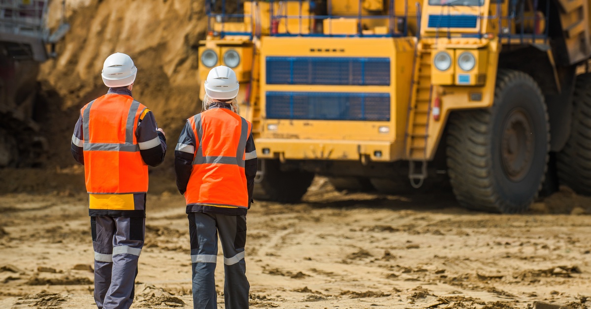 Two mining workers wearing helmets and high-visibility vests survey a mining site and mining vehicle.