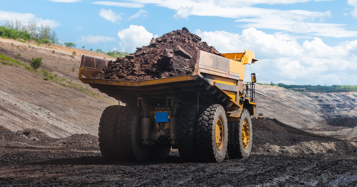A yellow dump truck hauls a large load of mined material through a muddy mining site. There are clouds in the sky.