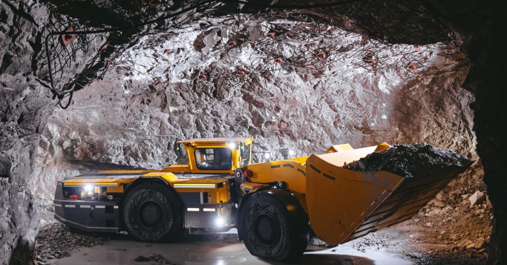 A large yellow mining vehicle is hauling a large load of mined material through a cavernous tunnel.