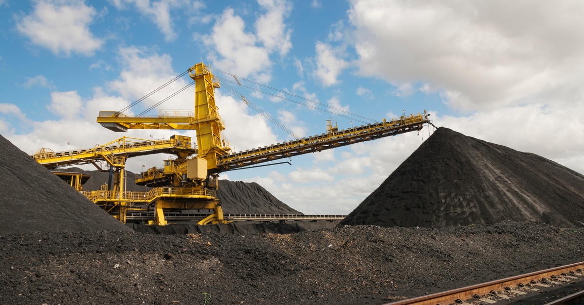 A mining site has a conveyor belt with coal going down the line while there's other large mining equipment in the background.