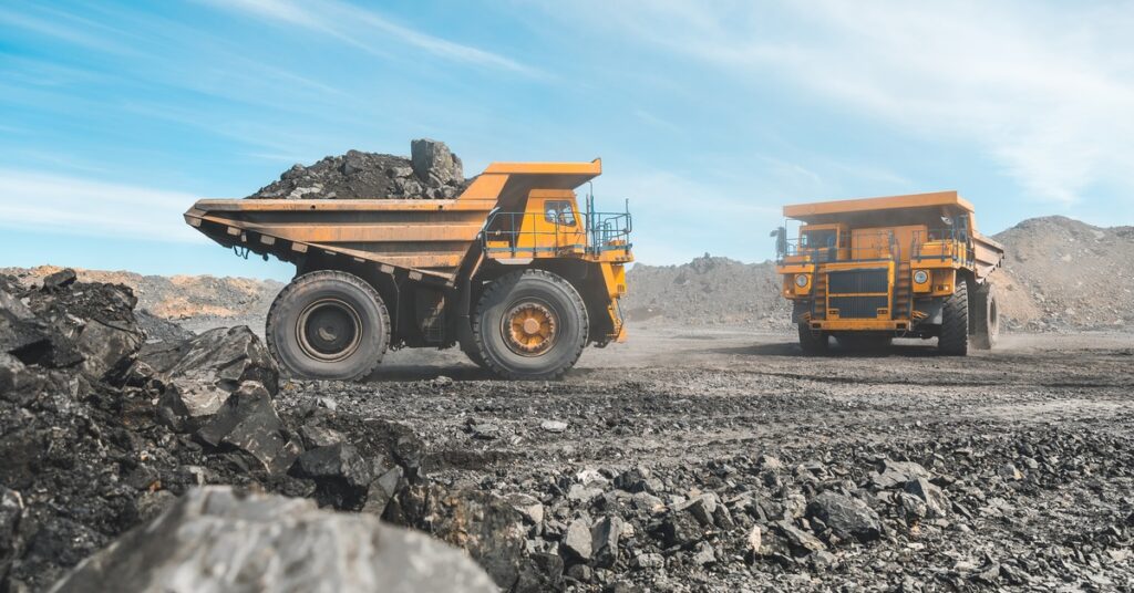 Two yellow mining vehicles drive around at a mining quarry, hauling different loads of raw ore with clouds in the background.