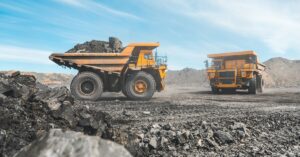 Two yellow mining vehicles drive around at a mining quarry, hauling different loads of raw ore with clouds in the background.